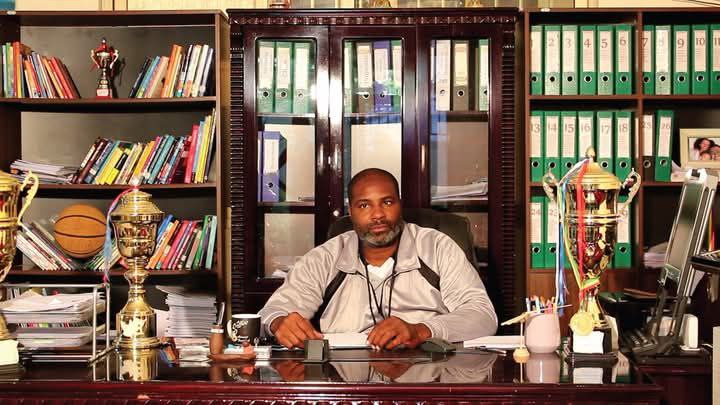 Coach Carlos seated in his office with basketball trophies and project records